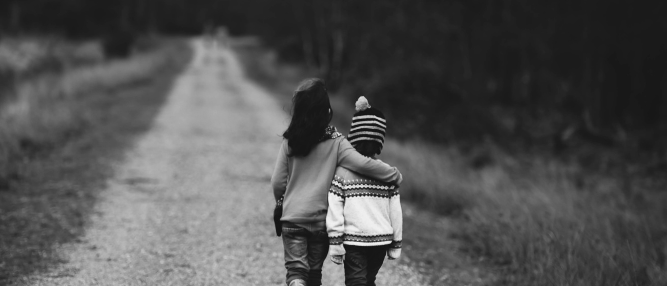 Black and white photo of two children walking along an unpaved road, one child has their arm over the other child