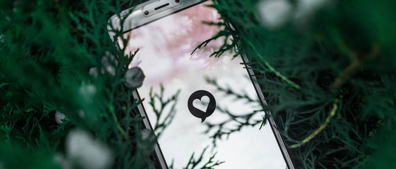 A cell phone with an image of a heart in a text bubble inside of a tree greenery surrounding it