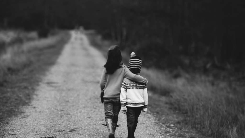 Black and white photo of two children walking along an unpaved road, one child has their arm over the other child